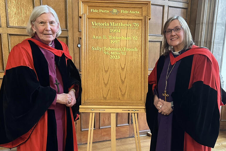 Bishop Victoria Matthews and Bishop Sally Johnson French line up with the Trinity College honours board featuring their names.